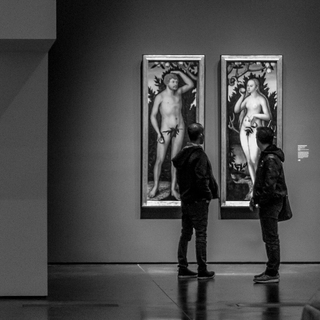 Photo of men standing in a gallery looking at nude art. The image is titled